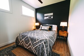 Un dormitorio con cama, dos mesitas de noche, una mesita auxiliar, una ventana y un cuadro colgado en la pared.