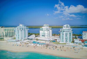 Un hotel frente a la playa con varios edificios de gran altura, un área de piscina y una playa con tumbonas.
