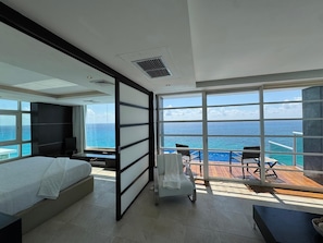 A modern hotel room with a large bed, a seating area with a view of the ocean, and a balcony with a wooden deck.