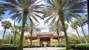 A hotel entrance with palm trees, a central flower bed, and a clear sky. - 3012- Great Family Home near Disney (private pool) (Kissimmee)