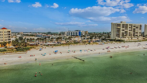 Ein Strand mit klarem türkisblauem Wasser, einem sandigen Ufer und Gebäuden im Hintergrund.