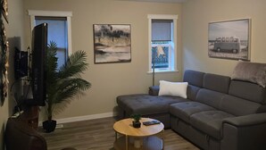 A living room with a sectional sofa, a round coffee table, a television, and two windows with artwork. - A Walk to the Harborfront  (Nanaimo)