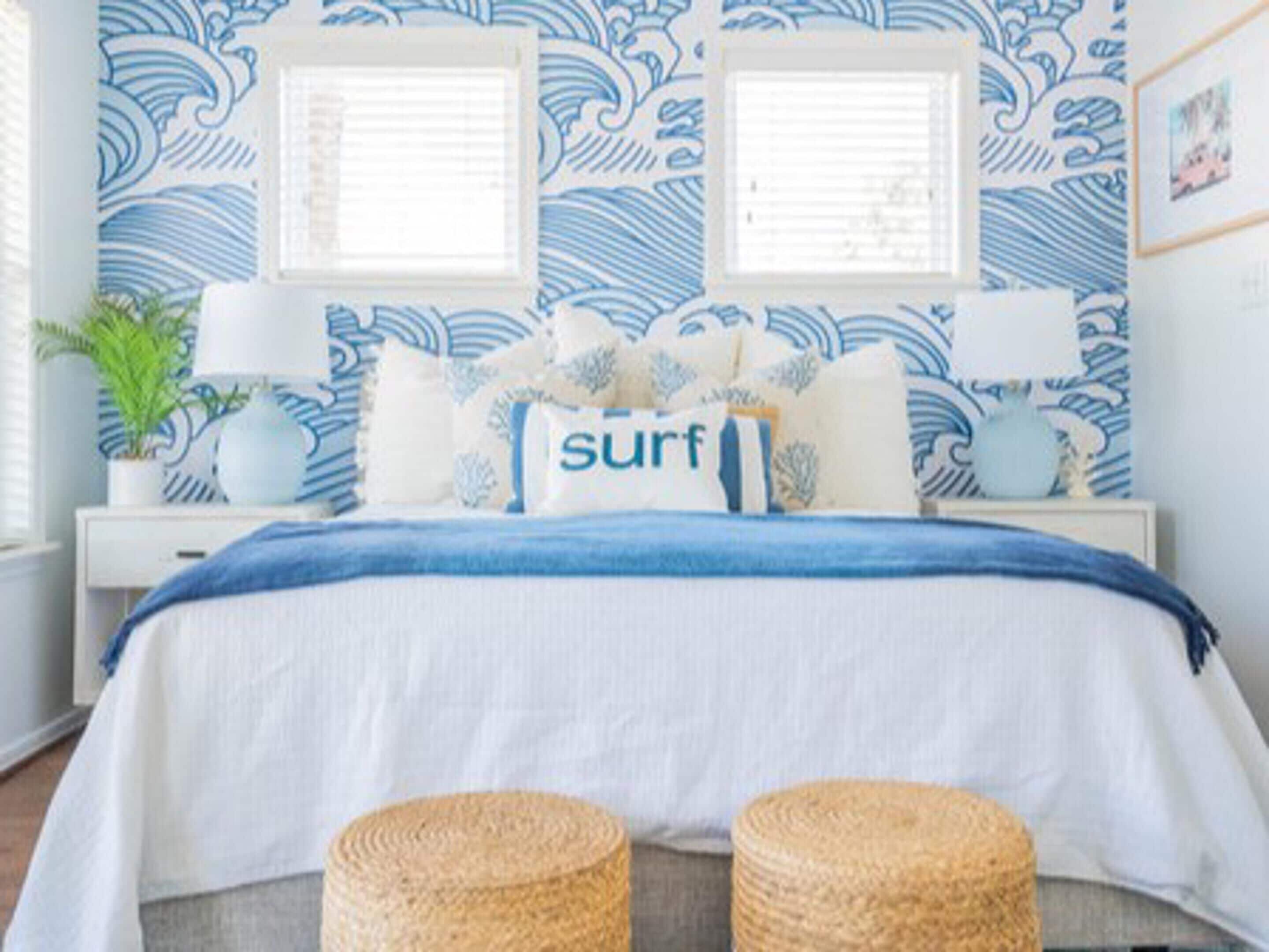 A bedroom with a blue and white wave-patterned wallpaper, a bed with a blue and white striped bedspread, two bedside lamps, and a potted plant.