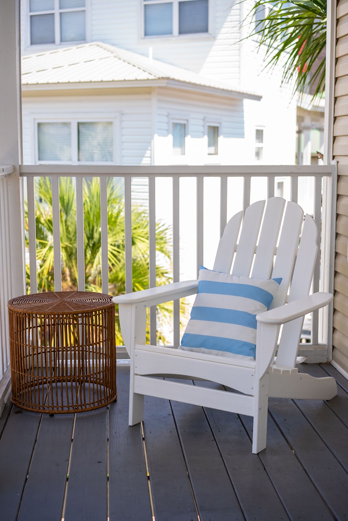 A white Adirondack chair with a blue and white striped pillow on a wooden deck, a wicker basket, and a view of a white building with windows.