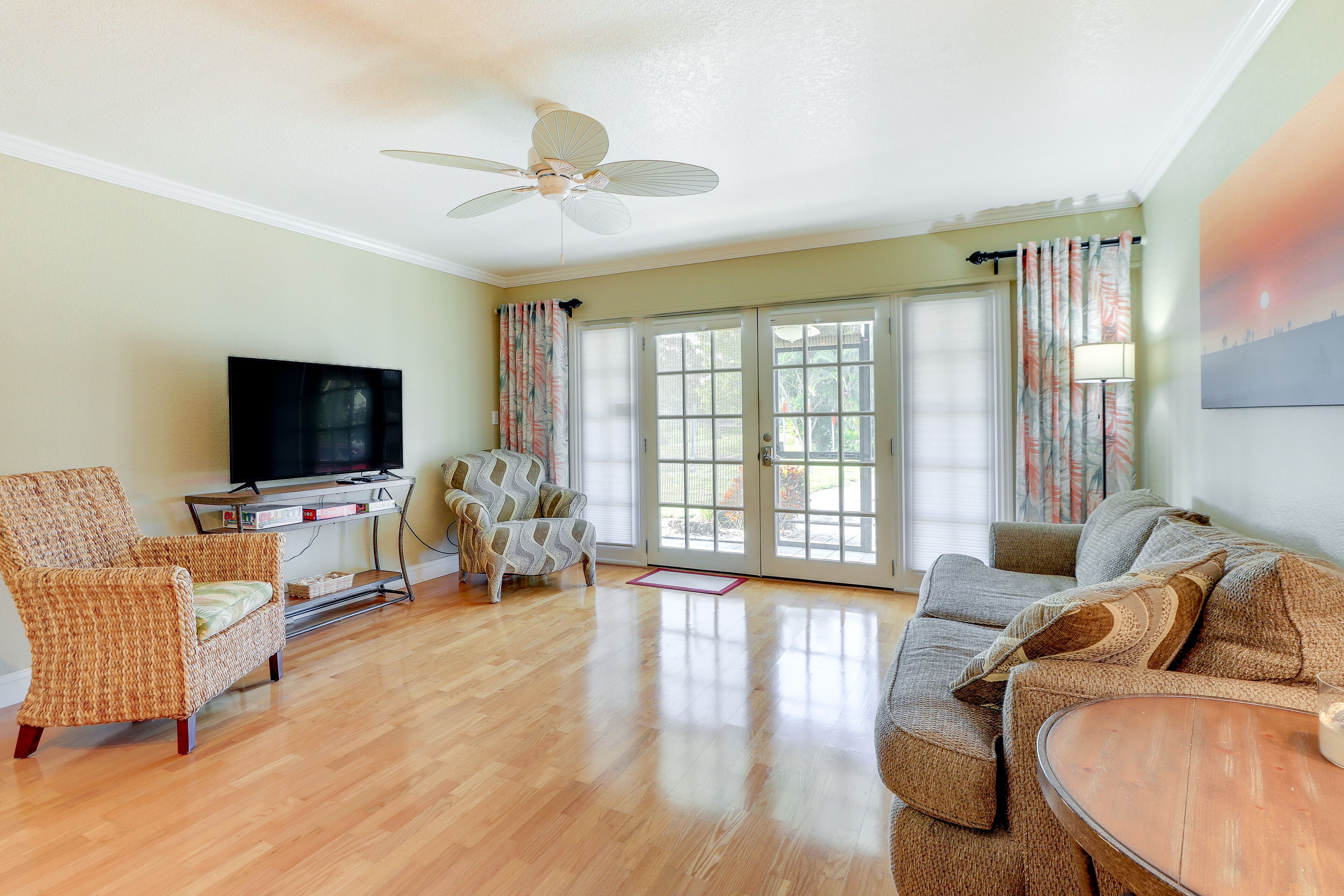 A living room with a television, two armchairs, a sofa, a ceiling fan, and a large window with curtains.