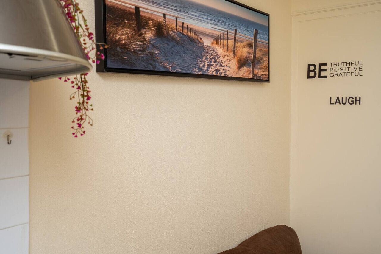 Een muur met een ingelijste foto van een strand, een bord met motiverende teksten en een kleine plant met roze bloemen.