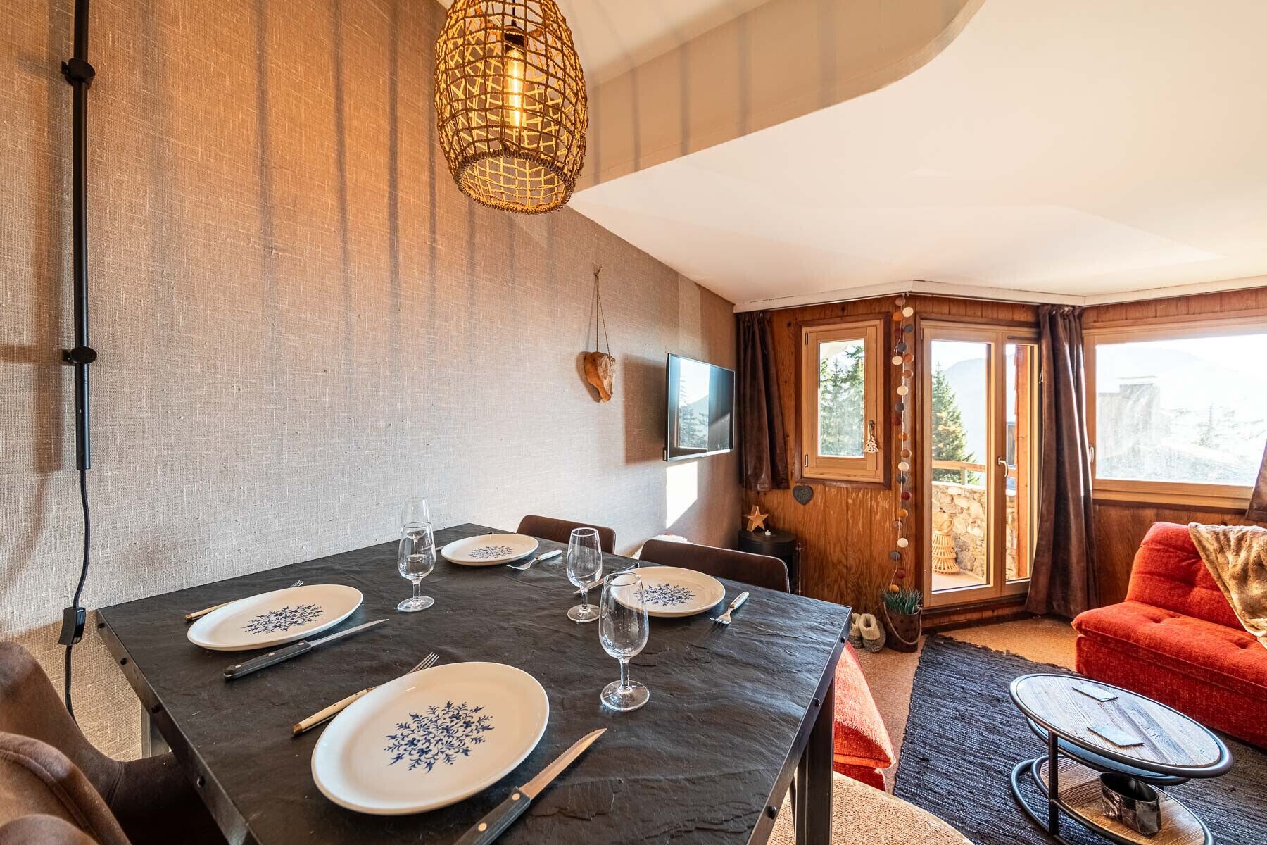 A dining area with a table set for four, a television on the wall, and a view of the outdoors through a glass door.