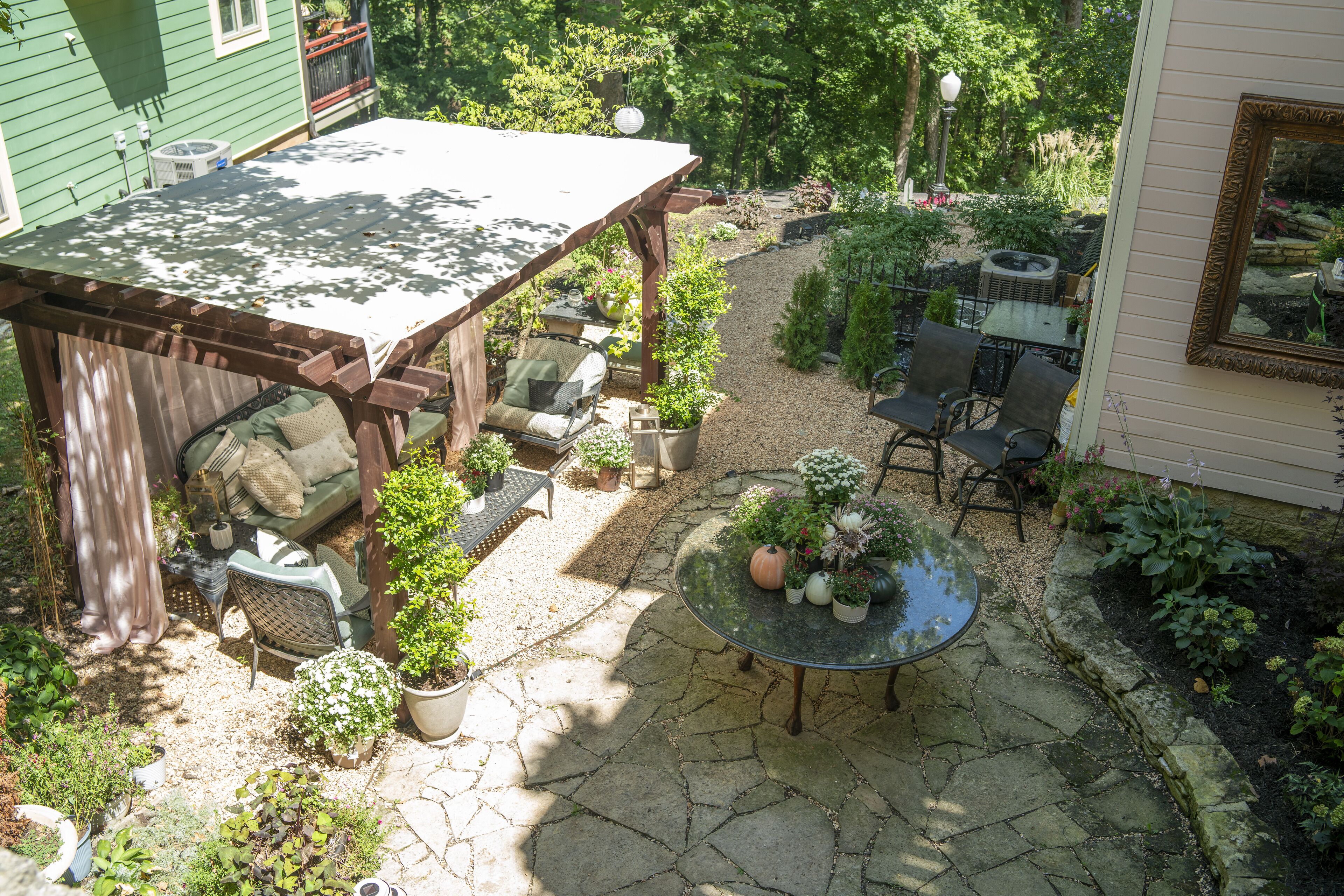 A patio with a wooden pergola, a round glass table, and several chairs. There are potted plants and a small garden area.