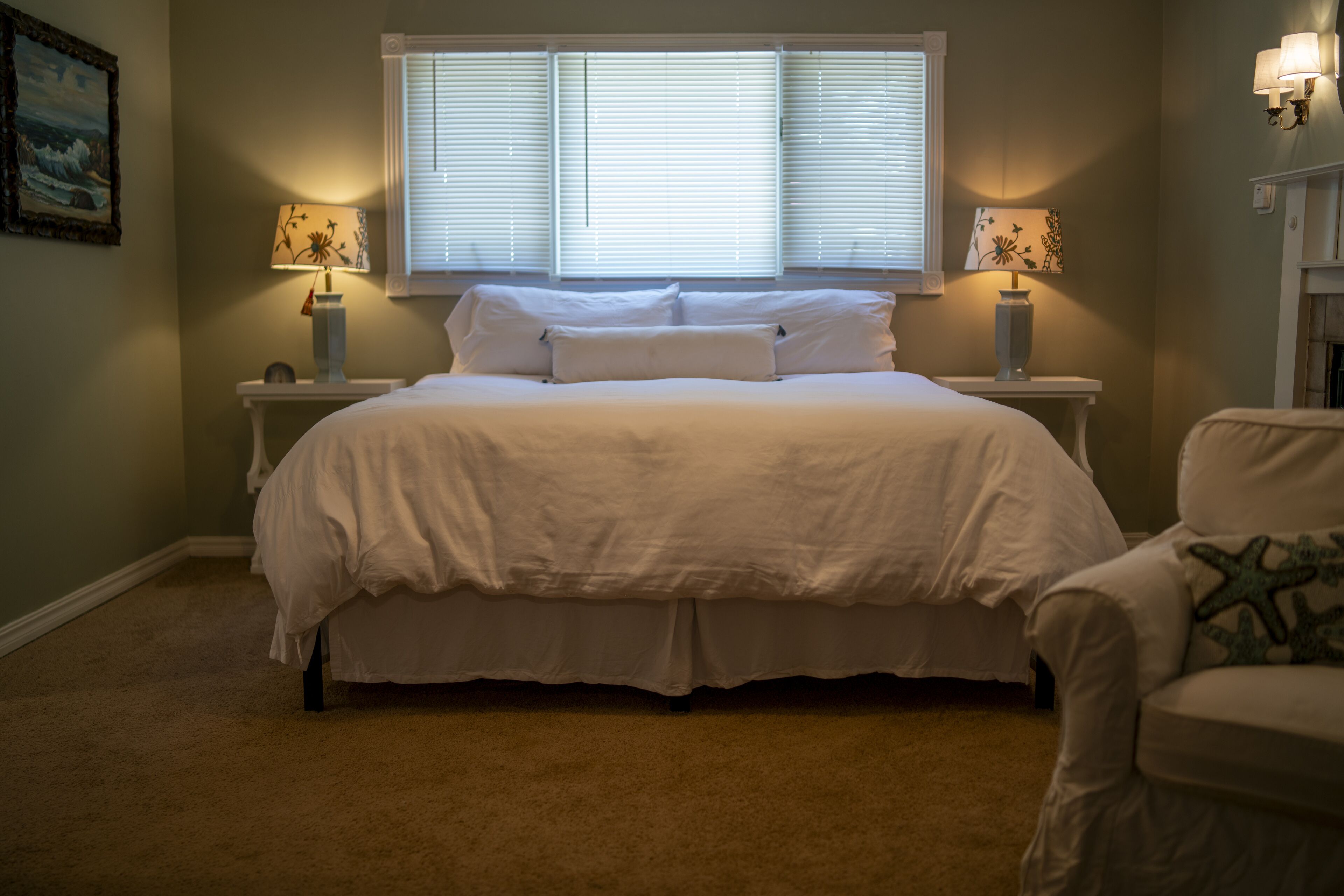 A bedroom with a large bed, two bedside tables with lamps, a chair, and a painting on the wall.