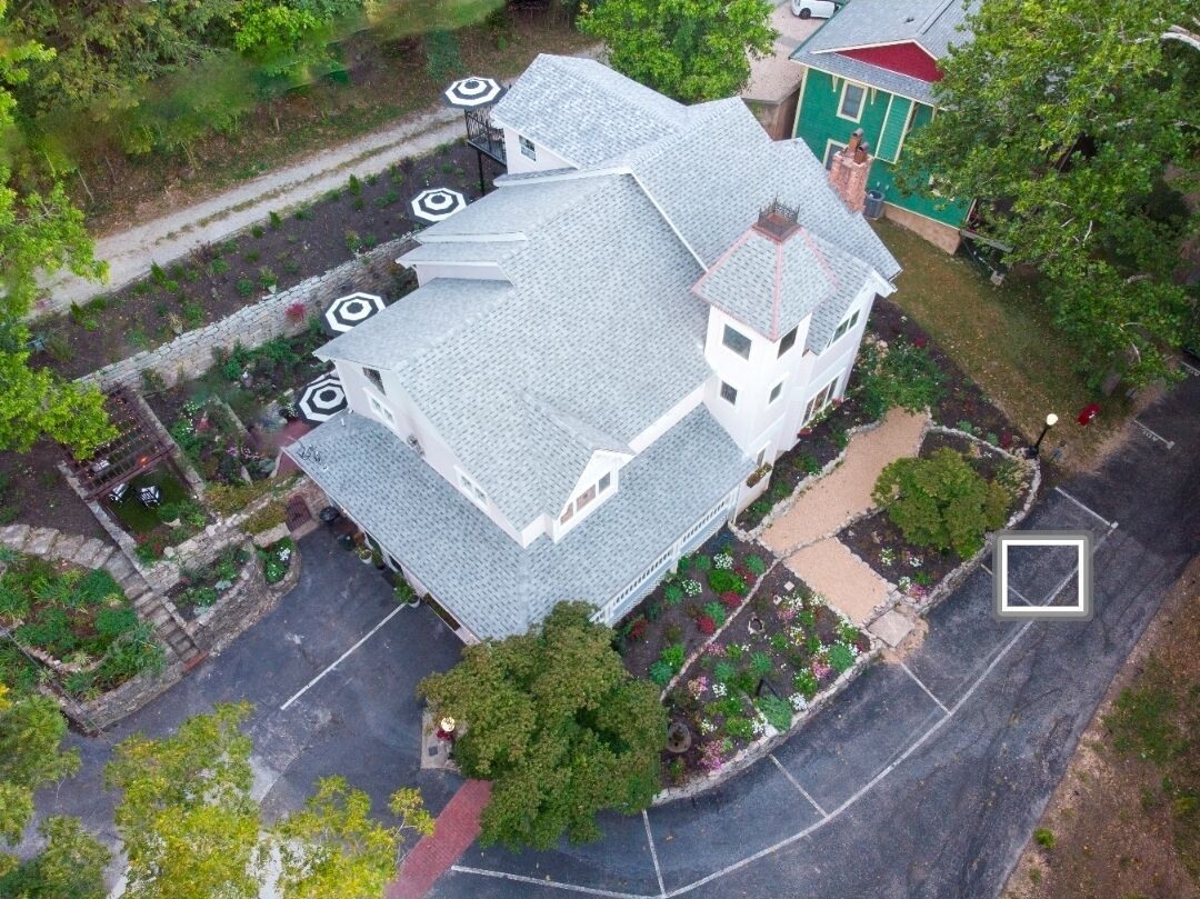 An aerial view of a building with a grey roof, surrounded by a landscaped garden with various plants and a paved driveway.