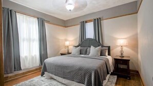 Un dormitorio con cama, dos mesitas de noche con lámparas, un ventilador de techo y ventanas grandes con cortinas.
