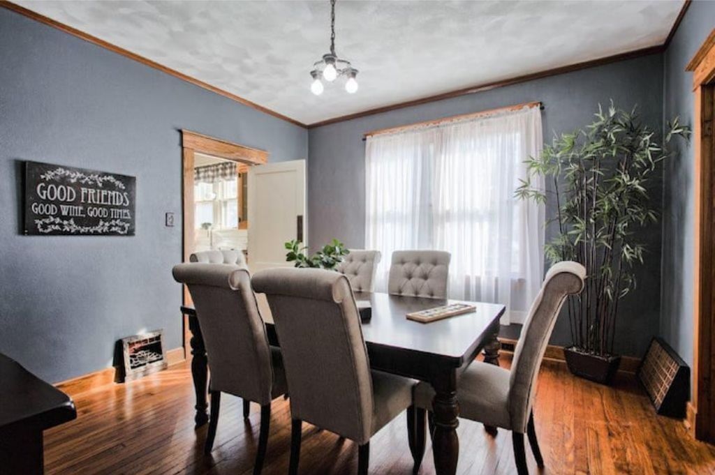 A dining room with a wooden table and four chairs, a mirror, a potted plant, and a sign on the wall.
