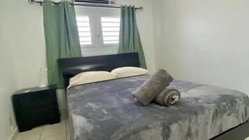 Un lit simple avec une couverture grise et deux oreillers, une table de chevet noire, une valise noire et une fenêtre avec des rideaux verts.
