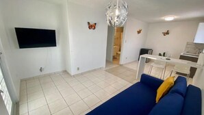 A living room with a blue sofa, a flat-screen TV mounted on the wall, a chandelier, and a kitchen area with a dining table and chairs. - Las Mariposas Suite Apt. 1 (Carolina)
