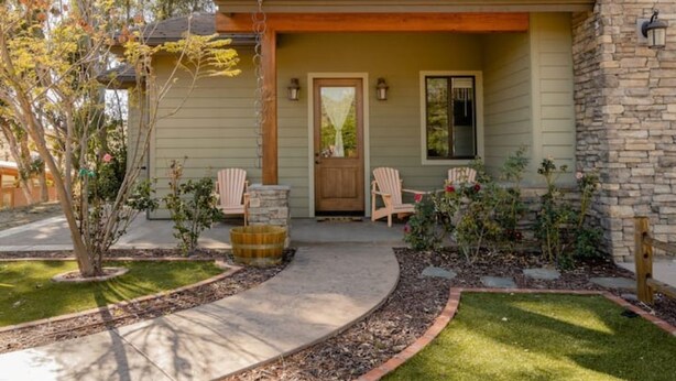 A house with a covered porch, two chairs, a wooden door, and a small garden with a tree and flowers.