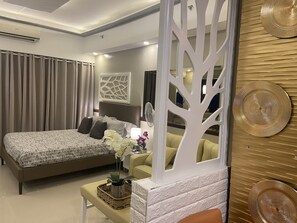 Une chambre moderne avec un grand lit, une table de chevet avec une lampe, un mur décoratif représentant des arbres et une vue sur l’intérieur de la pièce.