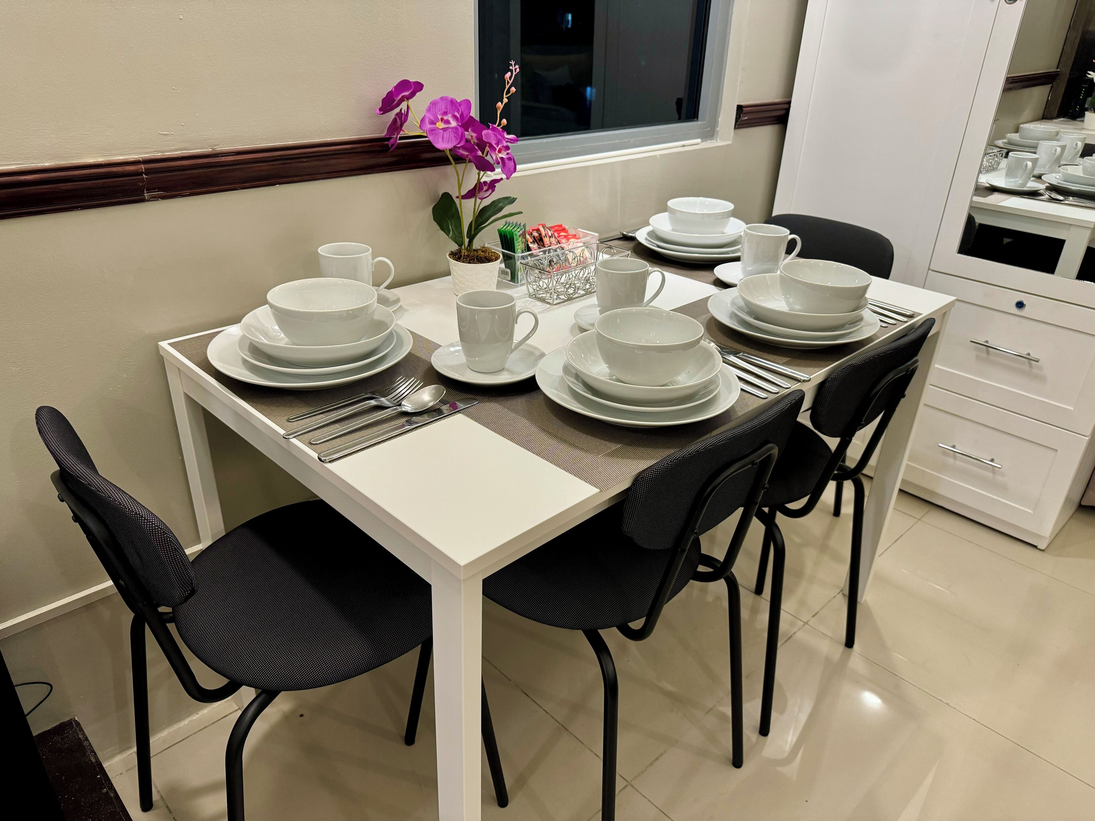 A dining table set with white plates, bowls, and cups, surrounded by black chairs.