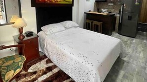 A bedroom with a bed, a desk, a chair, a hanging toy, and a kitchen area with cabinets and a refrigerator. - Pretty & Modern Private Studio 202 (Cali)