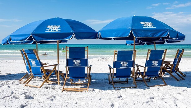 A beach with four blue beach chairs and two blue umbrellas with the Coral Reef Club logo.