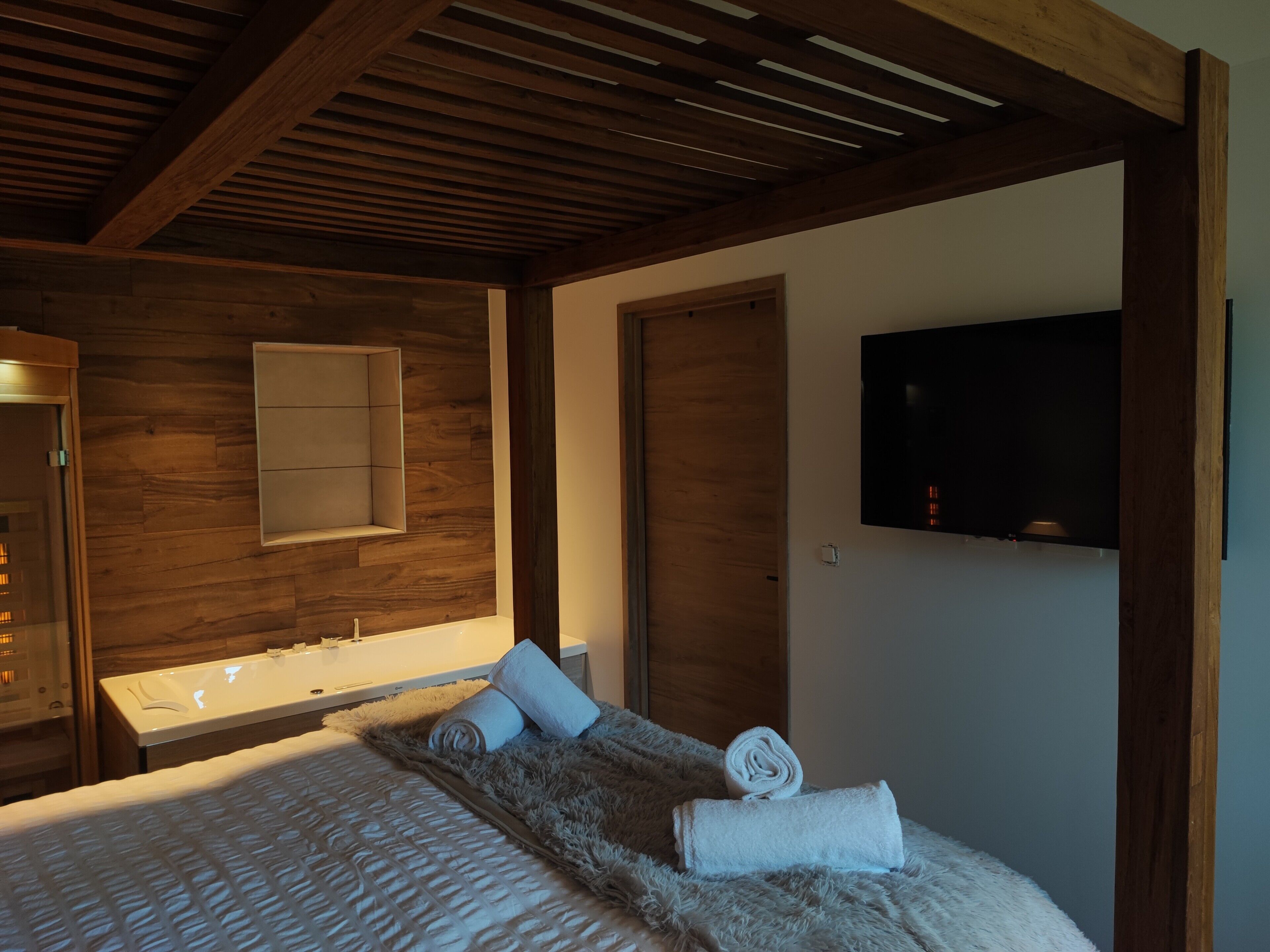 A modern hotel room with a wooden ceiling, a large bed with a fluffy blanket, a built-in bathtub, a flat-screen TV mounted on the wall, and a wooden door.