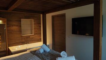 A modern hotel room with a wooden ceiling, a large bed with a fluffy blanket, a built-in bathtub, a flat-screen TV mounted on the wall, and a wooden door.