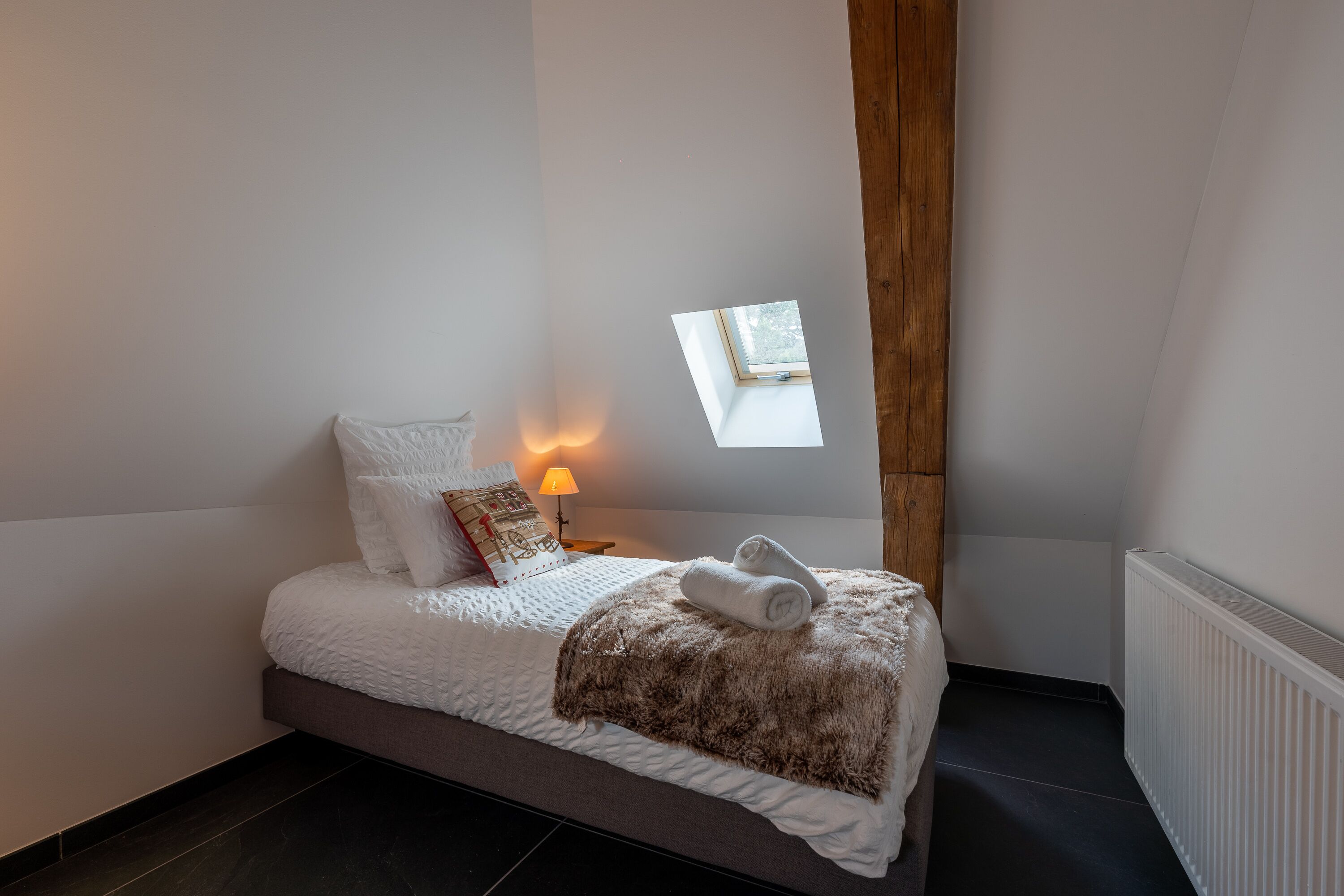 A bedroom with a bed, a nightstand, a lamp, a window, and a radiator.