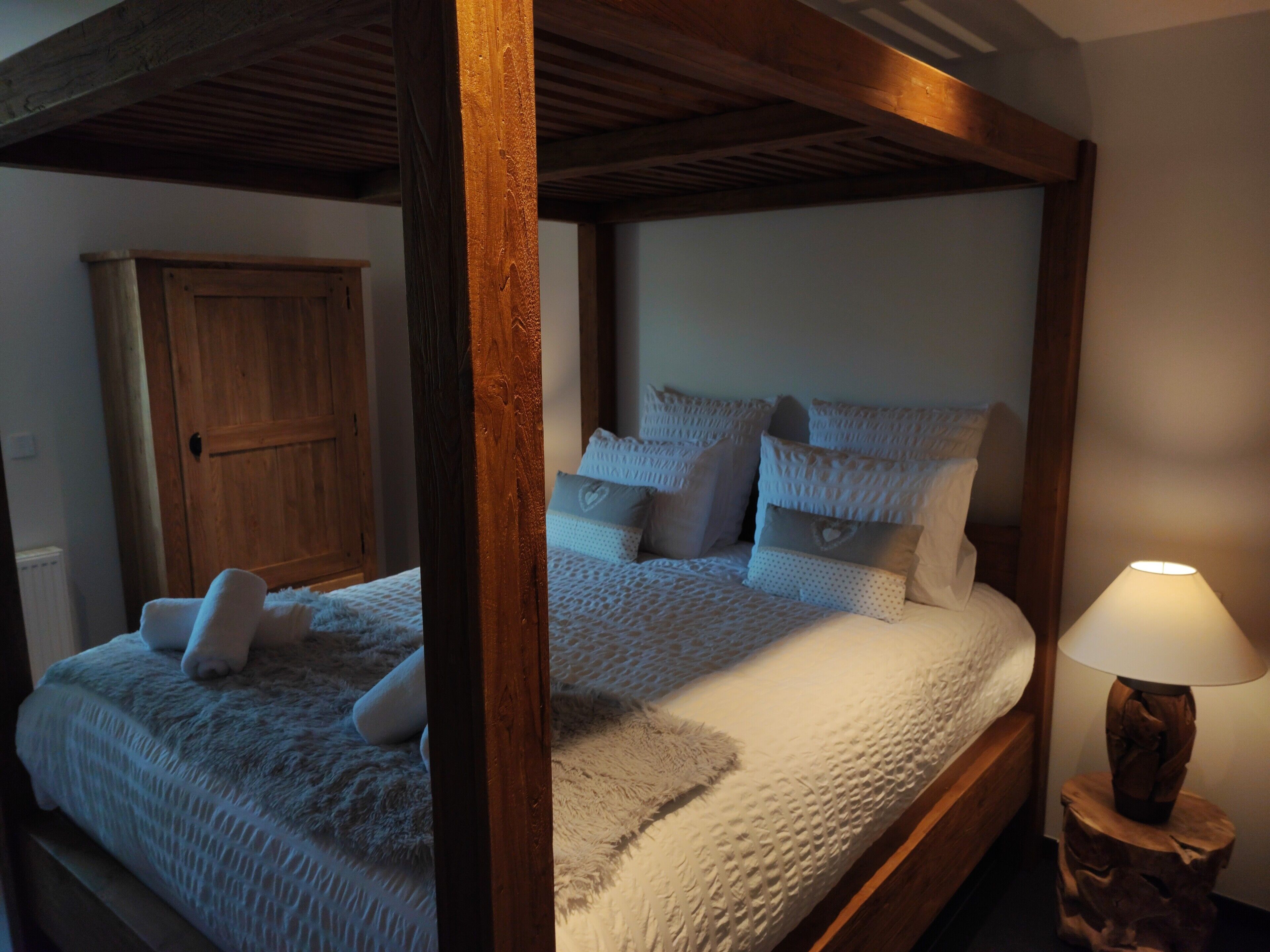 Een hemelbed met houten frame, witte beddengoed en meerdere kussens. Een houten nachtkastje met een lamp staat naast het bed.