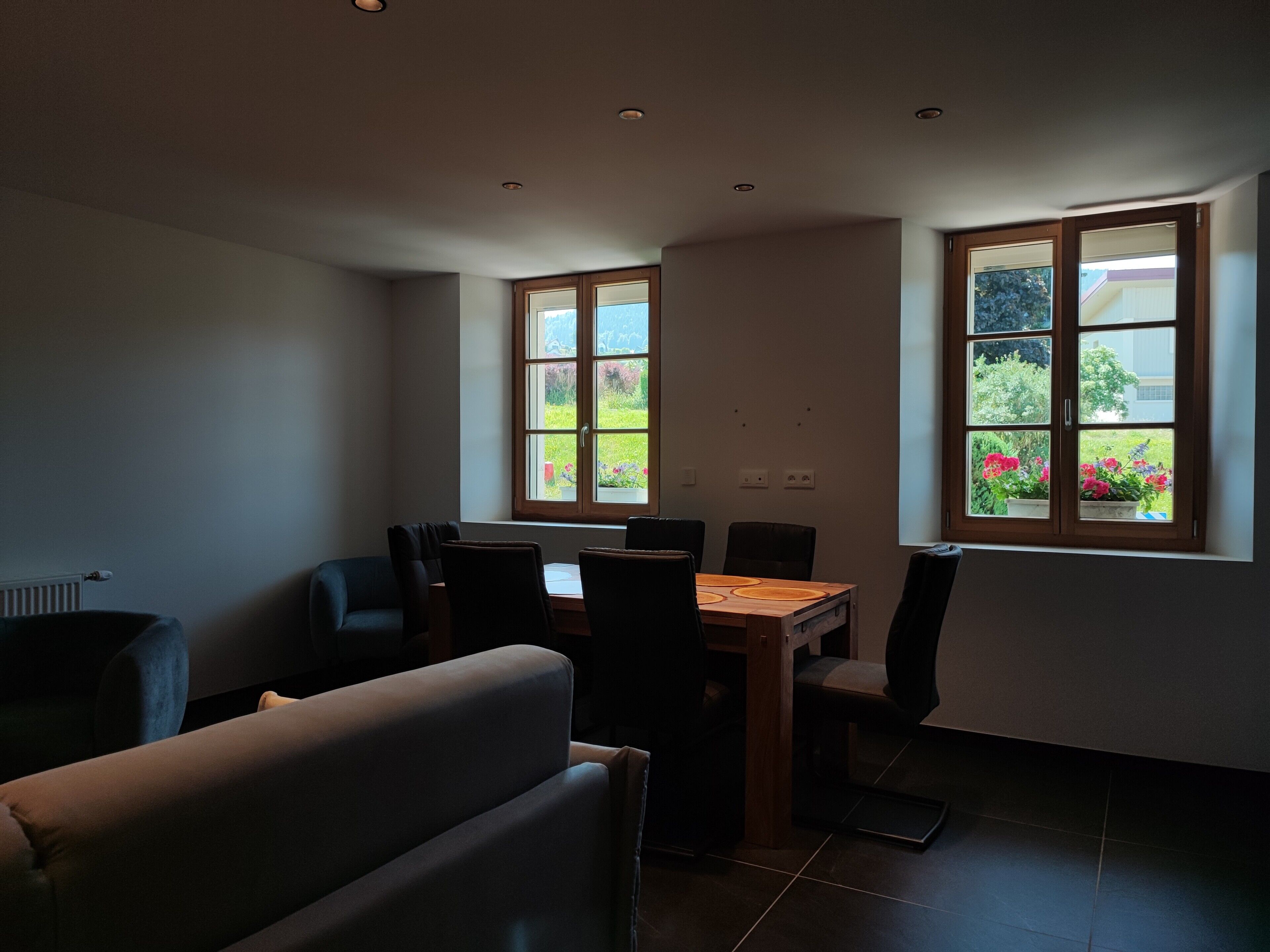 A dining area with a table and chairs, a sofa, and large windows with a view of greenery.