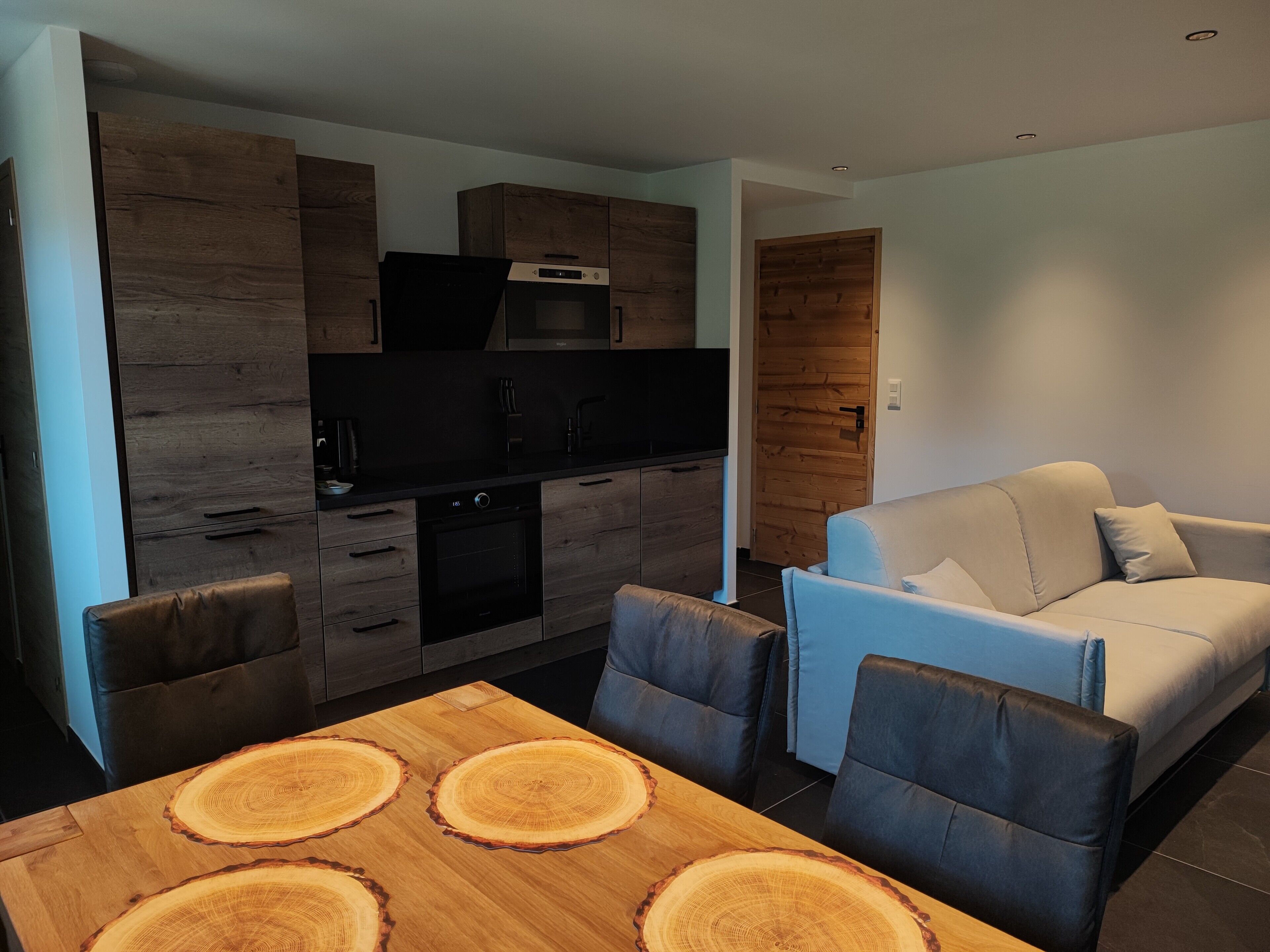 Una cocina moderna con armarios oscuros y un microondas integrado, una mesa de comedor con un centro de mesa de madera, y un sofá de color claro en la sala de estar.