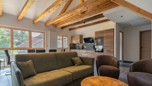 A modern living room with a sofa, chairs, and a coffee table. A kitchen is visible in the background with wooden cabinets and appliances.