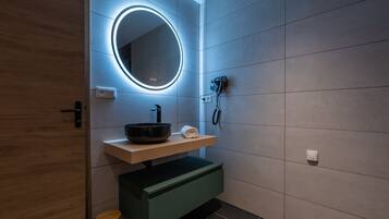 Un baño moderno con puerta de madera, un espejo redondo con iluminación indirecta, un lavabo negro, una toalla y un mueble de baño verde.