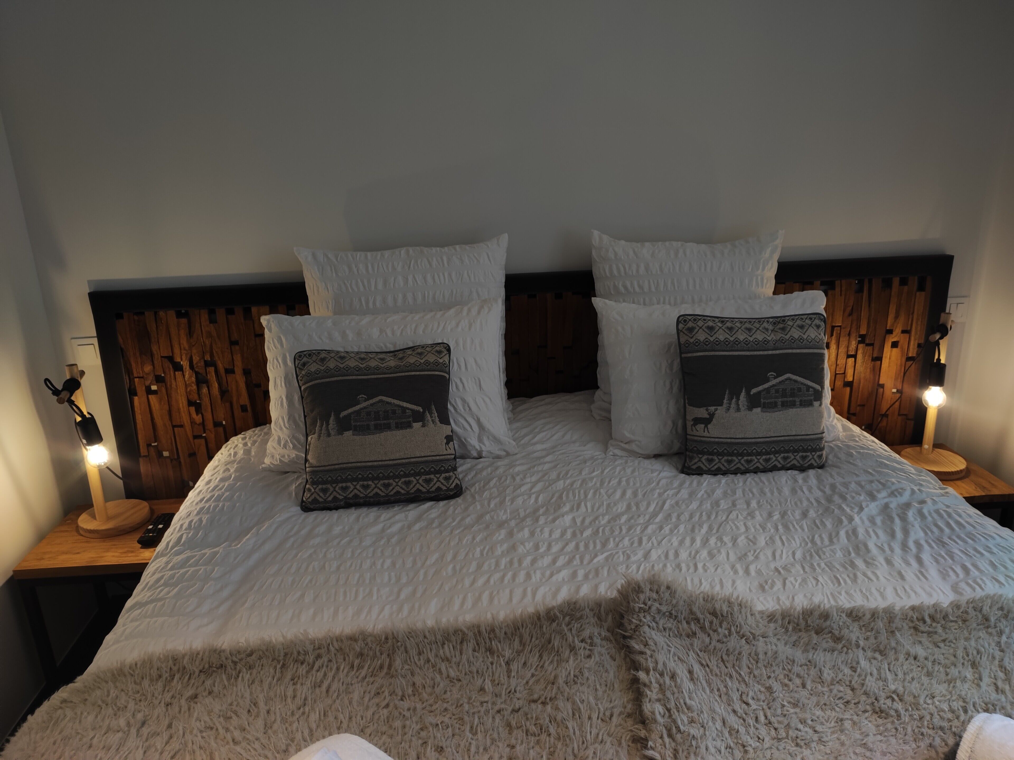 Ein ordentlich bezogenes Bett mit weißer Bettwäsche und zwei Zierkissen, ein Holz-Kopfteil und Nachttischlampen.