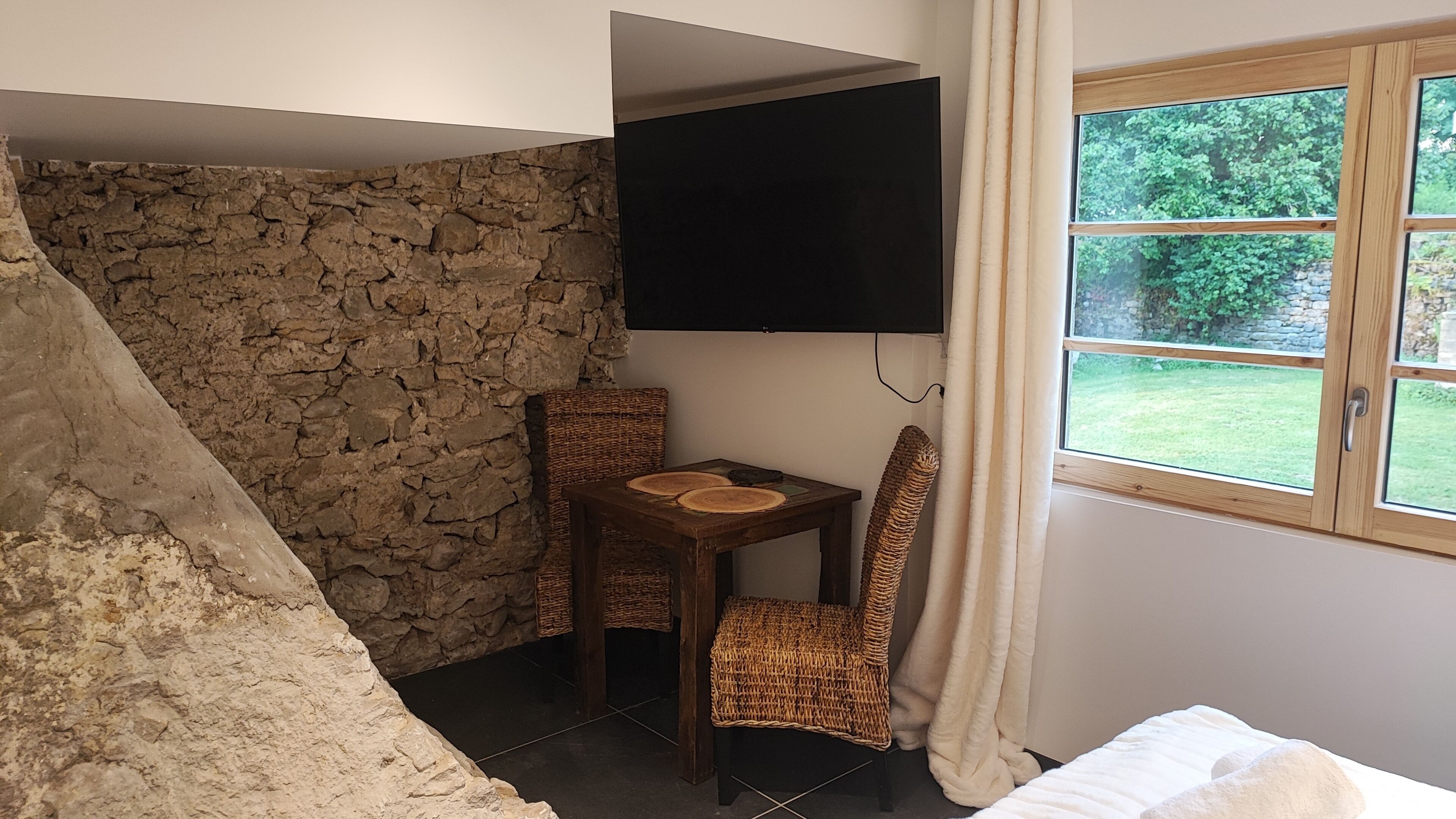 Een kamer met een stenen muur, een flatscreen televisie aan de muur, een klein tafeltje met twee geweven onderzetters, een rieten stoel en een raam met gordijnen.
