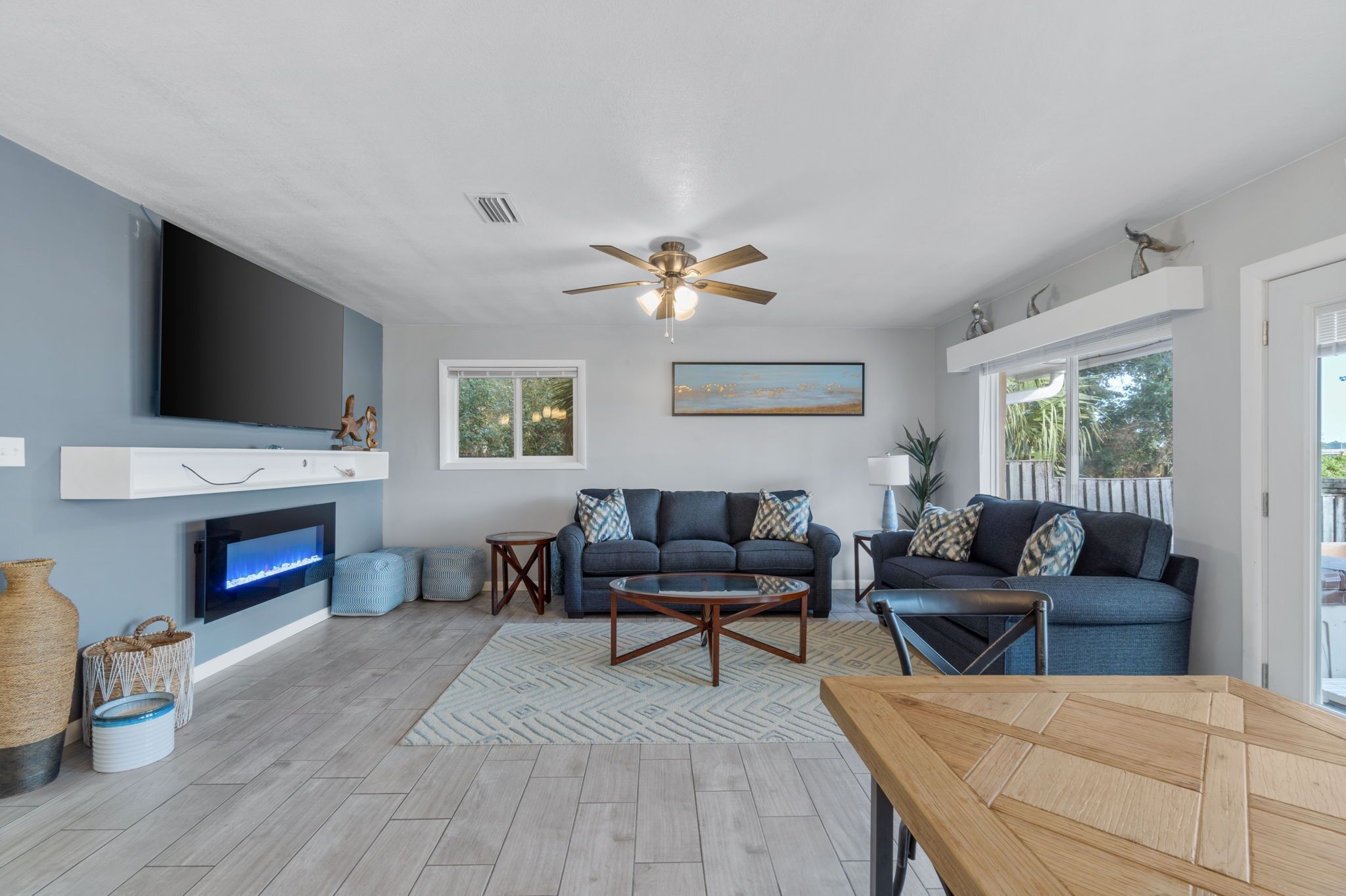 A modern living room with a flat-screen TV, ceiling fan, and a large window providing natural light.