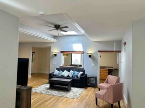 Un salon avec un canapé de couleur foncée, une table basse et un tapis à motifs. On y trouve un ventilateur de plafond, un luminaire fixé au mur et une fenêtre laissant entrer la lumière naturelle.