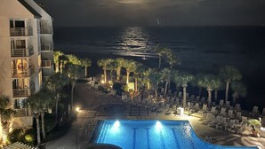 Una vista notturna di una zona piscina di un hotel con vista sull'oceano, palme e un edificio con balconi.