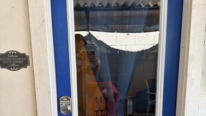 Una puerta azul con un panel de vidrio, un candado con teclado numérico y un letrero que dice 'Welcome Please Remove Your Shoes'.