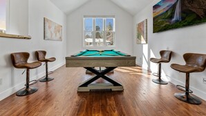 A pool table in a room with wooden flooring, white walls, and a large window.