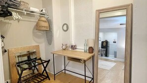 A small, well-lit room with a wooden floor, a desk with a mirror, a shelf with folded clothes, and a door leading to another room.