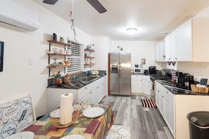 A compact kitchen with white cabinets, a stainless steel refrigerator, and a microwave. There are shelves with various kitchen items and a small dining area with a table set for two.