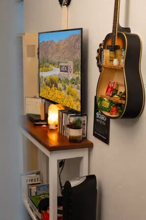 A room with a television displaying a landscape, a guitar, and a shelf with books and a plant.