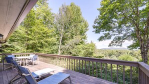Une terrasse en bois, dotée de chaises longues et d’une table, qui offre une vue sur une zone boisée, avec des arbres et des collines au loin.