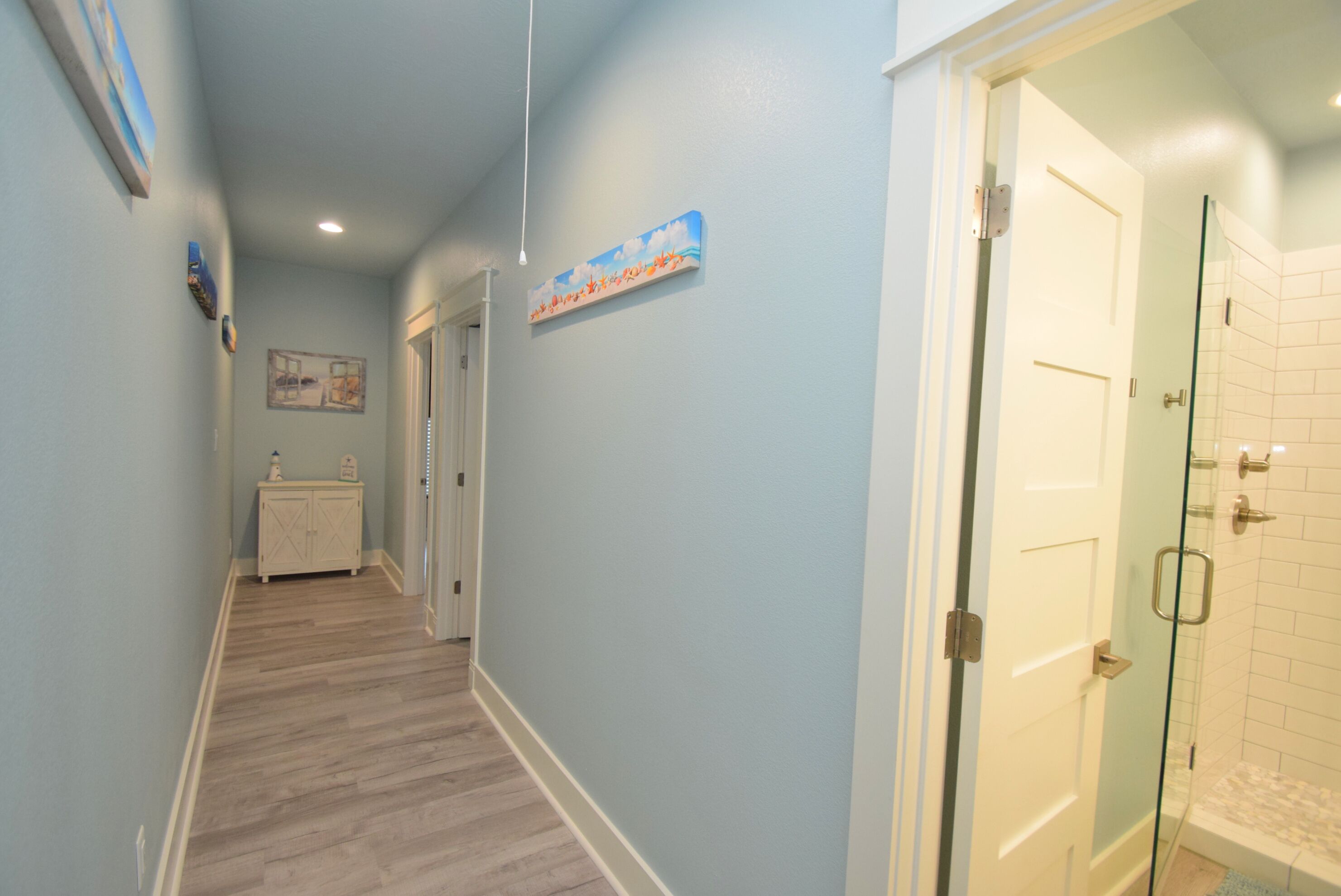 A hallway with light blue walls, wooden flooring, and a glass shower door leading to a bathroom.