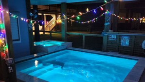 Ein Schwimmbecken mit Holzterrasse und einer Treppe, die zu einer erhöhten Ebene führt. Über dem Pool hängen bunte Lichterketten.