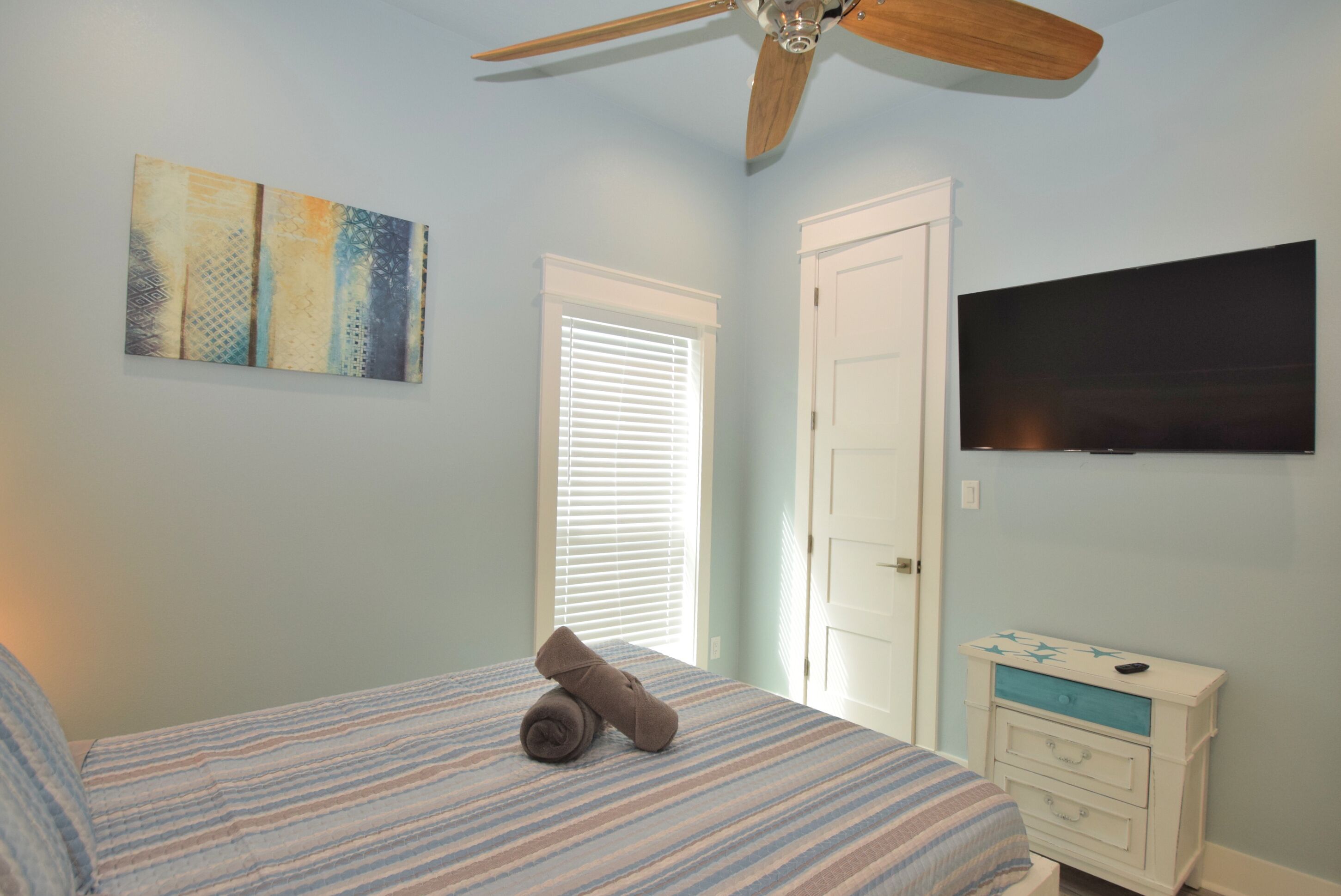 A bedroom with a bed, a ceiling fan, a window with blinds, a door, a dresser, and a TV mounted on the wall.