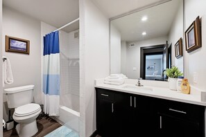 A modern bathroom with a white toilet, a shower with a blue and white curtain, a sink with a mirror, and a plant on the counter. -  North Campus (UT-Austin) Apt 7 /Red River/Moody Center (Austin)