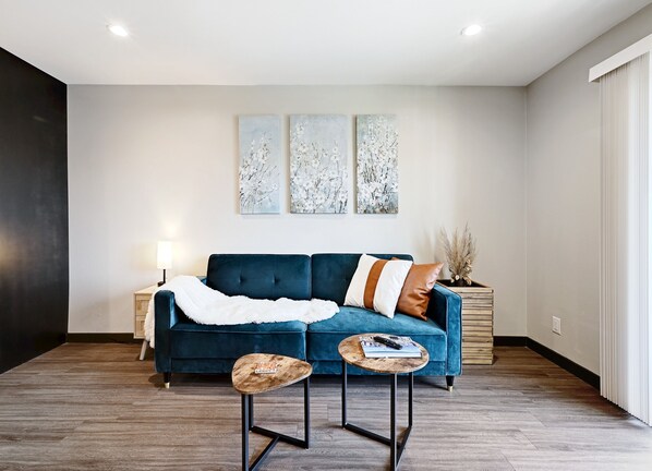 A modern living room with a blue sofa, a small wooden coffee table, and three abstract paintings on the wall. -  North Campus (UT-Austin) Apt 7 /Red River/Moody Center (Austin)