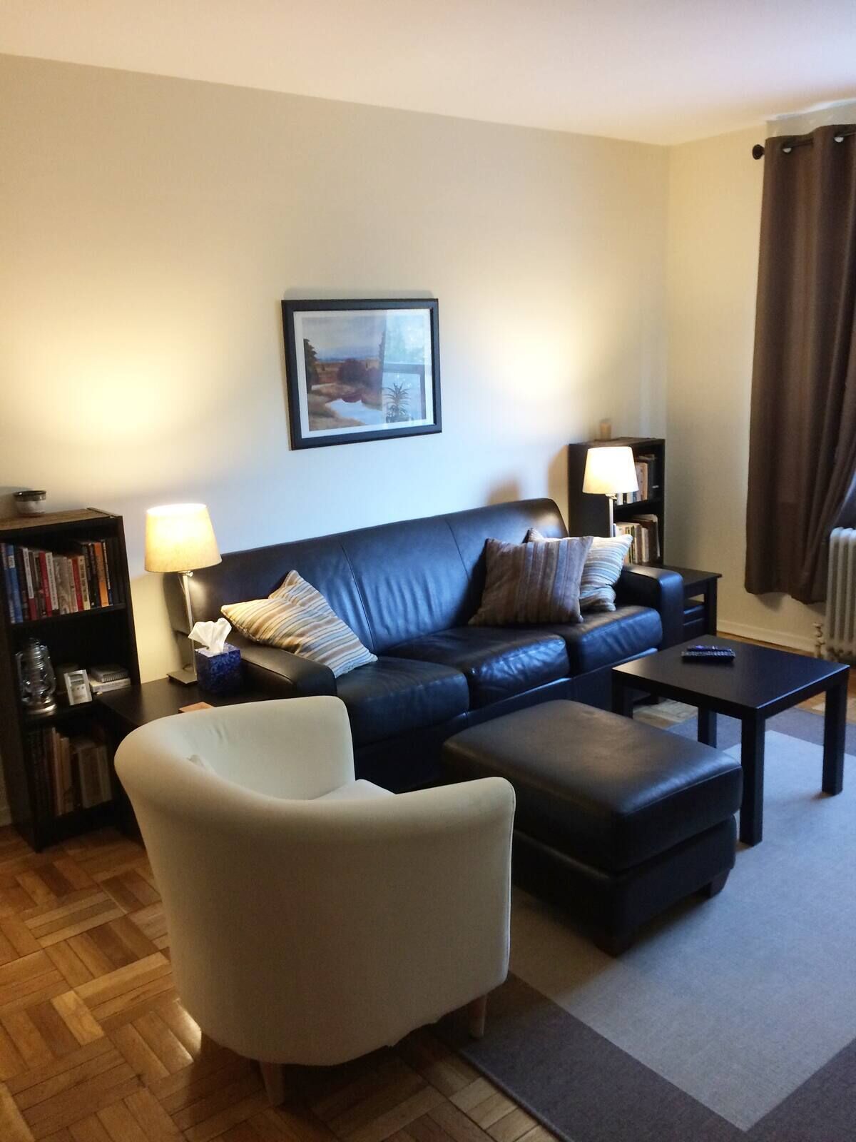 Ein Wohnzimmer mit einem dunkelblauen Sofa, einem weißen Sessel, einem Couchtisch, einem Bücherregal und einem gerahmten Bild an der Wand.