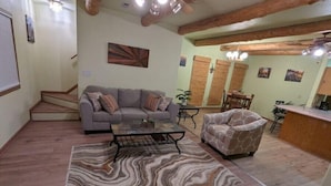 A living room with a sofa, armchair, coffee table, and a patterned rug. There is a staircase leading to another level, and a kitchen area with a counter and chairs. - Inviting & Spacious! Unique indoor and outdoor vibe! (Walsenburg)