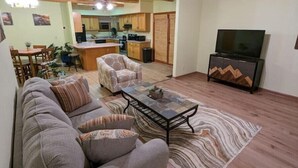 A living room with a sofa, armchair, coffee table, and a TV stand. There is a kitchen in the background with wooden cabinets and a dining area with a table and chairs. - Inviting & Spacious! Unique indoor and outdoor vibe! (Walsenburg)